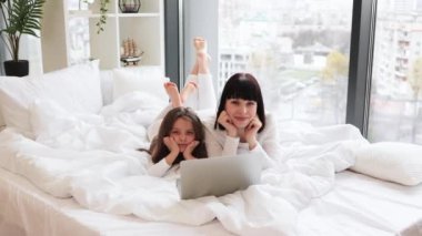 Anne ve kızı birlikte dizüstü bilgisayar kullanarak yatakta yatıyorlar. Konforlu ev ortamında mutlu aile bağları. Gülen yüzler sevgiyi, birlikteliği ve rahatlığı gösterir..