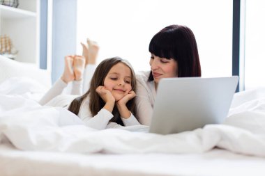 Anne ve kızı dizüstü bilgisayar kullanarak yatakta yatıyor. Çocuk ekranda gülümseyen elleri üzerinde dinleniyor. Yatak odasında teknolojiyle aile bağı kurma anı. Mutlu ve rahat bir atmosfer.