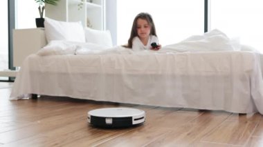 Modern bir yatak odasında beyaz yatakta uzanmış, tahta zeminde robot elektrik süpürgesini gözlemleyen bir kız. Minimalist dekorlu ve nötr renklere sahip bir oda.