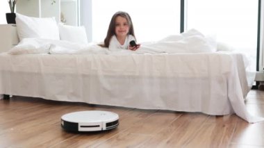 Yatakta dinlenen genç bir kız parke zemin temizleme odasında robotik elektrik süpürgesi izliyor. Modern teknoloji, otomasyon ve ev işleri için kolaylık.
