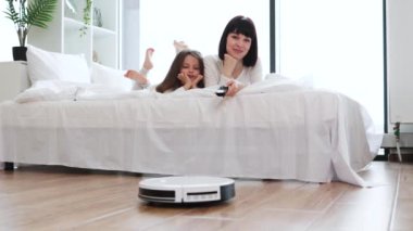 Anne ve kızı yatağa uzanıp robot elektrikli süpürge izliyorlar. Aile, ahşap döşemeleri ve beyaz yatakları olan temiz, modern bir evde eğleniyor..