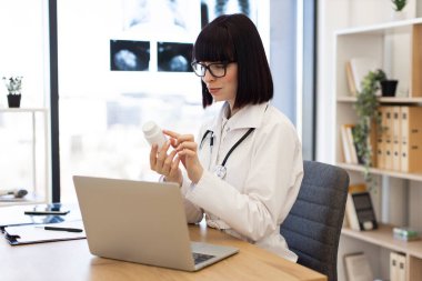 Gözlüklü ve laboratuvar önlüklü bir kadın doktor masada dizüstü bilgisayarla oturuyor. Doktor ilaç şişesini inceliyor. Arka planda dokümanlar ve raflar olan modern tıp bürosu.