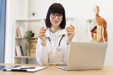 Gözlüklü ve steteskoplu bir kadın doktor tıbbi ofisteki masasında hapları tutuyor, güvenle gülümsüyor. Modern tıp uzmanları sağlık hizmetleri tavsiyesi veriyor.