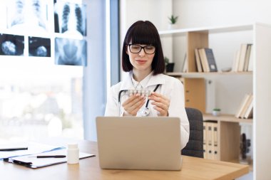 Modern tıp bürosunda dizüstü bilgisayarda çalışan gözlüklü kadın doktor elinde bir paket hap tutuyor. Ofiste tıbbi çizelgeler, raflarda kitaplar ve teşhis ekipmanları yer alıyor..