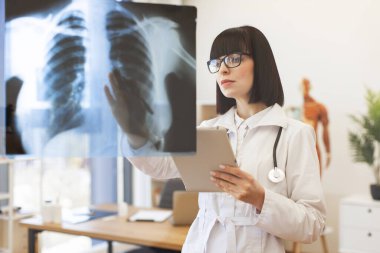 Kadın doktor modern tıp ofisindeki cam panoda göğüs röntgenini analiz ediyor. Teşhis için odaklanmış profesyonelliği ve teknolojinin kullanımını gösteriyor. Sağlık radyolojisi ve tıbbi uzmanlık kavramı.