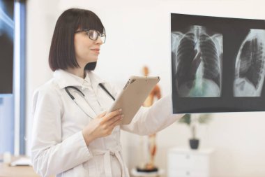 Kadın doktor cam panoda hastanın röntgenlerini inceliyor. Tıp uzmanı laboratuvar önlüğü ve steteskop giyer, teşhis görüntülerini analiz eder. Olay yeri hastane içinde sağlık hizmetleri veya klinik ortamlar öneriyor.