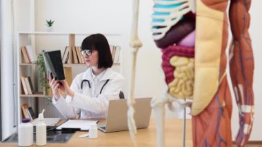 Modern tıp ofisinde röntgen analizi yapan kadın doktor. Beyaz önlük ve gözlük takan tıp uzmanı hasta teşhisi üzerinde çalışıyor. Odada anatomik model, kitaplıklar ve masa var..