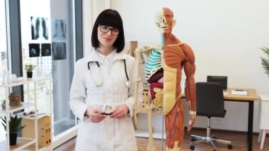 Tıp ofisinde anatomik modelin yanında duran doktor steteskop ve gözlük takıyor. Sağlık ve eğitim kavramlarının vurgulanması ve tıbbi ekipmanlarla donatılmış parlak bir ortam.