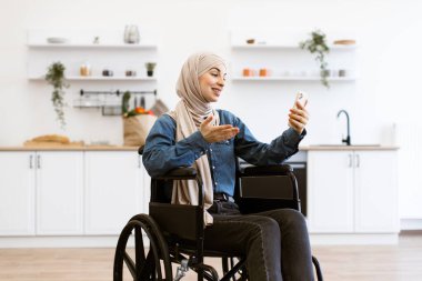 Modern ev mutfağında akıllı telefon kullanarak tekerlekli sandalyedeki gülümseyen Müslüman kadın. Engellilik, teknoloji ve bağımsızlık kavramı. Tekerlekli sandalye kullanıcısı tesettüre bağlanıyor.