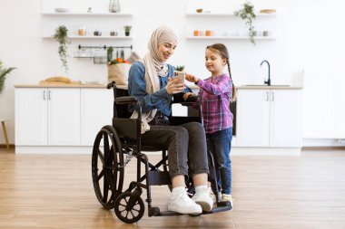 Anne ve kızı teknolojinin tadını çıkarıyorlar, bağlanma anları yaratıyorlar. Tekerlekli sandalyedeki Müslüman anne ve genç kızı modern mutfakta akıllı telefon kullanıyorlar..