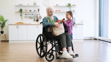 Tekerlekli sandalyedeki anne ve kızı ellerinde kese kağıdıyla modern mutfağın arka planında taze sebzeler tutuyor. Resim, anne ve çocuk arasındaki aile, destek ve birlikteliği çağrıştırıyor, bağ vurgulanıyor.