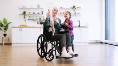 Tekerlekli sandalyedeki Müslüman anne ve modern mutfakta selfie çeken kız. Akıllı telefondan çekilen mutlu aile anı. Anne başörtüsü takıyor, çocuk ona sarılıyor..