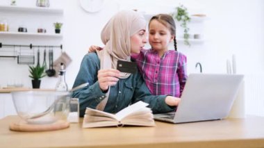 İnternetten alışveriş yaparak aile bağı kurmak ve mutfakta yemek pişirmek. Müslüman anne ve kız mutfak masasında oturup yemek malzemeleri alıyorlar dizüstü bilgisayar, banka kartı ve yemek tarifi kitabı kullanarak..