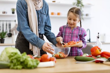 Parlak mutfak ortamındaki aile bağları sağlıklı yemek ve yemek pişirmeyi teşvik ediyor. Müslüman anne kız salata kasesine tahtadan kesilmiş sebze dökün..