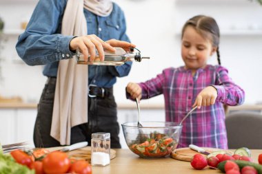 Müslüman anne ve kız mutfakta yemek hazırlarken salata kâsesine zeytinyağı ekliyor. Birlikte yemek pişirmek aile bağlarını ve sağlıklı yeme alışkanlıklarını geliştirir. Aşçılık becerilerini yakalar.