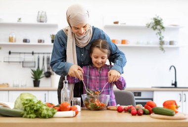 Aile bağı ve sağlıklı beslenme kavramı. Müslüman anne ve kızı, parlak mutfakta salata kaselerinde sebze karıştırıyorlar. Sağlıklı yemek hazırlarken gülümsüyor ve eğleniyoruz..