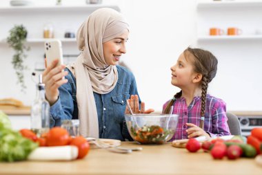 Müslüman anne ve kızı sağlıklı salata gösterirken görüntülü görüşme yapıyorlar. Mutfak masasında oturuyorlar, etrafları taze sebzelerle çevrili, gülümsüyorlar ve birlikte anın tadını çıkarıyorlar..