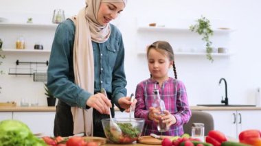 Modern mutfakta sağlıklı yemek pişirmek yerine aile bağlarını güçlendirmek. Müslüman anne ve kız salata kasesine zeytinyağı ekliyor. Taze sebzeler masada..