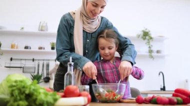 Aile bağı ve sağlıklı beslenme kavramı. Müslüman anne ve kızı, parlak mutfakta salata kaselerinde sebze karıştırıyorlar. Sağlıklı yemek hazırlarken gülümsüyor ve eğleniyoruz..