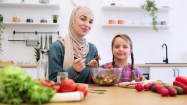 Müslüman anne ve kızı açık mutfak ortamında taze salatanın tadını çıkarıyorlar. Sağlıklı beslenmeye, aile bağlarına ve mutluluğa odaklanır..