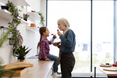 Müslüman anne ve kız modern mutfakta sağlıklı salatanın tadını çıkarıyorlar aile bağlarını ve sağlıklı yaşam tarzını sergiliyorlar. Anne başörtüsü takıyor, kız yemek yerken gülümsüyor..