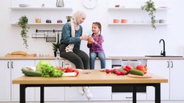 Türbanlı bir kadın, çevresi sebzelerle çevrili bir çocukla kaliteli vakit geçiriyor. Modern mutfak ortamında Müslüman anne ve kızı birlikte sağlıklı meyve suyu içiyor. Sağlıklı yaşam tarzı yerine aile bağı..