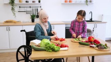 Tekerlekli sandalyedeki Müslüman anne ve kızı mutfakta salata için sebze kesiyor. Sağlıklı yemek yaparken kaliteli zaman geçirmek, aile bağları, dahil etme ve takım çalışması sergilemek.