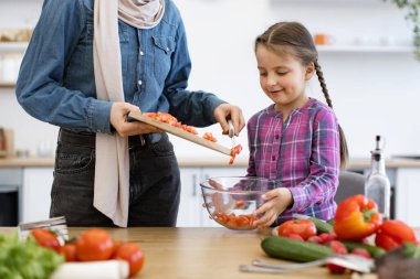 Mutfakta aile bağı. Müslüman anne ve kızı salata kasesine tahta tahtadan kesilmiş sebzeler döküyorlar. Sağlıklı yaşam tarzı ve besleyici yemek hazırlığı.