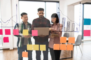 Veri analizi ve beyin fırtınası üzerine çalışan bir grup profesyonel iş toplantısına katılıyor. Takım renkli yapışkan notlarla saydam tahtadaki çizelgeleri tartışıyor.