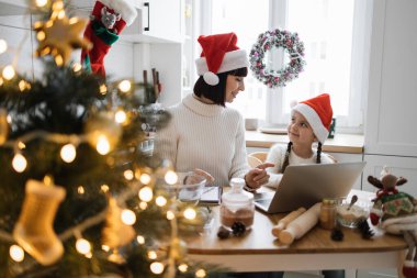 Noel Baba şapkalı beyaz anne ve kız Noel için dekore edilmiş sıcak mutfakta kurabiye pişiriyorlar. Tarif için dizüstü bilgisayar kullanıyorlar. Noel ağacı ve çelenkli ılık bir atmosfer..