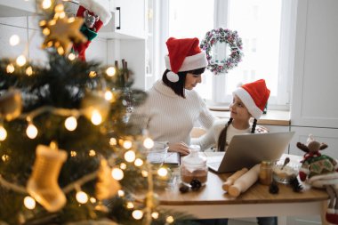 Kafkas anne ve kızı Noel boyunca mutfakta birlikte vakit geçiriyorlar. İkisi de Noel Baba şapkası takıyor, tatil hazırlıkları sırasında masada dizüstü bilgisayarla bağ kuruyor..