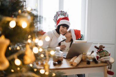 Kafkasyalı anne ve kız Noel Baba şapkası takıyor ve Noel 'in tadını çıkarıyor. Kadın ve kız, dizüstü bilgisayar ve dekore edilmiş ağacın yanındaki şenlikli anı paylaşıyor. Sıcak tatil atmosferi kapalı alanda yakalandı.