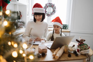 Kafkasyalı anne ve genç kız Noel Baba şapkası takıyor ve sıcak mutfakta birlikte kurabiye pişiriyorlar. Noel süslemeleriyle güçlendirilmiş şenlikli bir ortam. Annesinin yazdığı gibi dizüstü bilgisayarda çalışan odaklanmış çocuk..