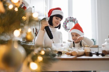Kafkasyalı anne ve genç çocuk mutfakta tatilin tadını çıkarıyor. İkisi de Noel Baba şapkası takıyor, malzemeleri elektrikli mikserle karıştırıyor. Ön plandaki Noel ağacı ışıkları sıcak şenlik ortamı yaratıyor.