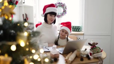 Gülümseyen beyaz anne ve kız Noel Baba şapkası takıyor, Noel 'in keyfini çıkarıyorlar. İkisi de masada dizüstü bilgisayarla otururken kameraya el sallıyor..