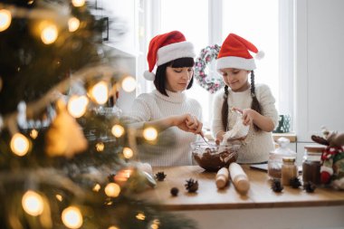 Asyalı bir kadın ve genç bir kız Noel Baba şapkası takıyor ve rahat bir mutfakta kurabiye pişiriyor. Noel ışıkları ve dekorasyonları şenlikli bir ortam yaratır..