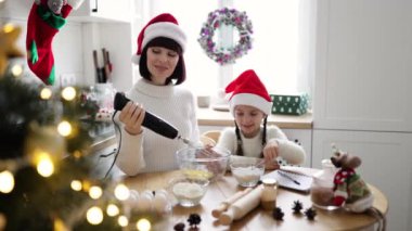 Kafkasyalı anne ve kız Noel Baba şapkası takıp birlikte kurabiye pişiriyorlar. Modern mutfakta Noel için dekore edilmiş malzemeleri karıştırıyorlar..