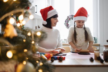 Kafkasyalı kadın ve genç kız Noel Baba şapkası takıp rahat mutfak ortamında kurabiye pişiriyorlar. Noel süslemeleri görünen şenlikli bir ortam.