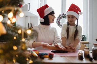 Genç beyaz anne ve kız Noel Baba şapkası takıp birlikte kurabiye pişiriyorlar. Noel ışıklarıyla süslenmiş rahat mutfakta şenlikli bir etkinliğin tadını çıkarıyorlar..