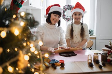 Evde Noel kurabiyesi pişirirken gülümseyen beyaz anne ve kız. Her ikisi de Noel Baba şapkası takıyor, neşeli bir ortam yaratıyor..