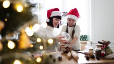 Kafkasyalı mutlu anne ve kız Noel Baba şapkası takıyor, hamuru sıcacık mutfakta yoğuruyor ve tatil sezonuna hazırlanıyor. Noel süslemeleri ile şenlikli bir ortam, ailede sevgi ve neşe göstergesi