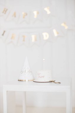 Pembe iki süslemeli ve uyumlu parti şapkalı beyaz pastanın olduğu doğum günü kutlaması sahnesi. Yumuşak renklerle minimalist ve zarif bir stil. İkinci yaş günü etkinlikleri ya da çocuk partileri için ideal.