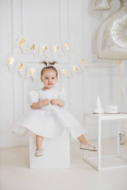 İkinci doğum günü kutlaması sırasında beyaz elbiseli, beyaz elbiseli genç beyaz bir bayan. Odada doğum günü afişi, balonlar ve pasta var..