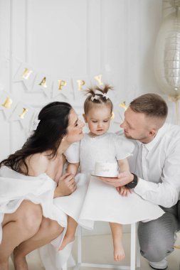 Beyaz anne, baba ve küçük kızı ikinci doğum gününü kutluyorlar. Ebeveynler, üzerinde iki tane yazılı küçük kek tutan çocuğa sevgiyle bakıyorlar. Aile bağları ve neşeli anlar.