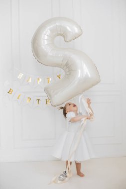 Beyaz odada büyük bir balon tutan tatlı bir kız, doğum gününü kutluyor. Beyaz elbiseli beyaz bir çocuk doğum günü afişinin yanında durmuş, neşeyle yukarı bakıyor..