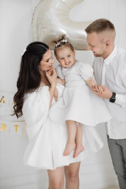 Kafkas ebeveynler ve küçük kızı doğum gününü aydınlık odada kutluyorlar. Erkek neşeyle eğlenirken, kadın kucağında bebek.