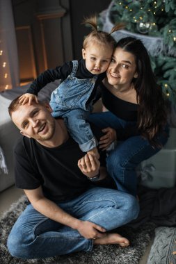 Yeni yürümeye başlayan bebekle birlikte genç beyaz ebeveynler Noel ağacının yanında oturmuş, mutlu bir şekilde gülümsüyorlar. Aile, tatil sezonunu rahat bir oturma odasında kutlar. Aile bağlarında sevgi ve sıcaklığı yakalamak.