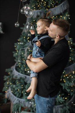 Beyaz bir baba Noel ağacını süslerken küçük kızını tutuyor. Süs eşyalarına uzanan çocuk neşeli ve şenlikli bir ortam yaratıyor..