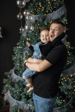 Kafkasyalı baba ve kızı, ışıklarla süslenmiş Noel ağacının yanında mutlu bir şekilde poz veriyorlar. Günlük kıyafetlerle gülümseyen adam kot pantolon giyen neşeli genç kızı tutuyor ve sıcak tatil ruhunu yakalıyor..