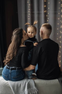 Caucasian mother kissing toddler daughter while father sits nearby. Family enjoying quality time together indoors, surrounded by soft lighting. Cozy and intimate family setting. clipart